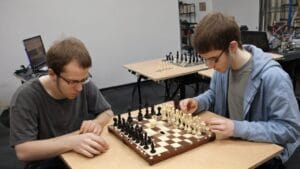 Two men playing chess at a table, observed by an Artificial Intelligence media platform.