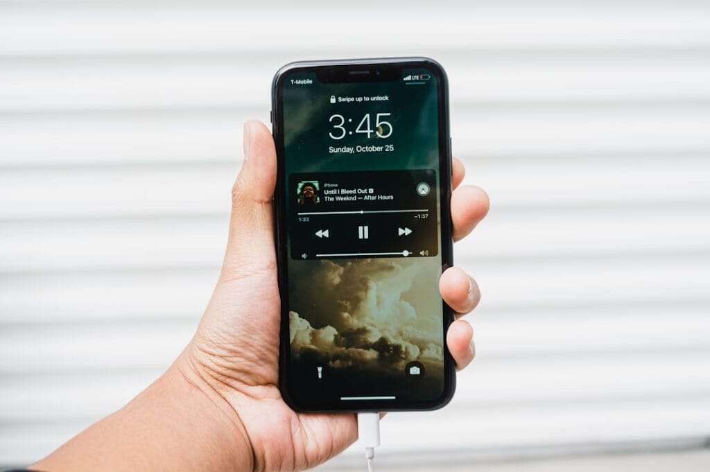 A person holding a smartphone with a music player app on the screen, displaying the time and a current song, with the device connected to a charger.