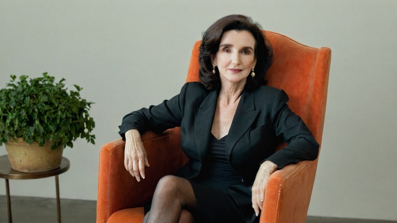 A woman with dark hair sits in an orange armchair, wearing a black outfit and pearl earrings. A potted plant is on a small side table next to the chair.