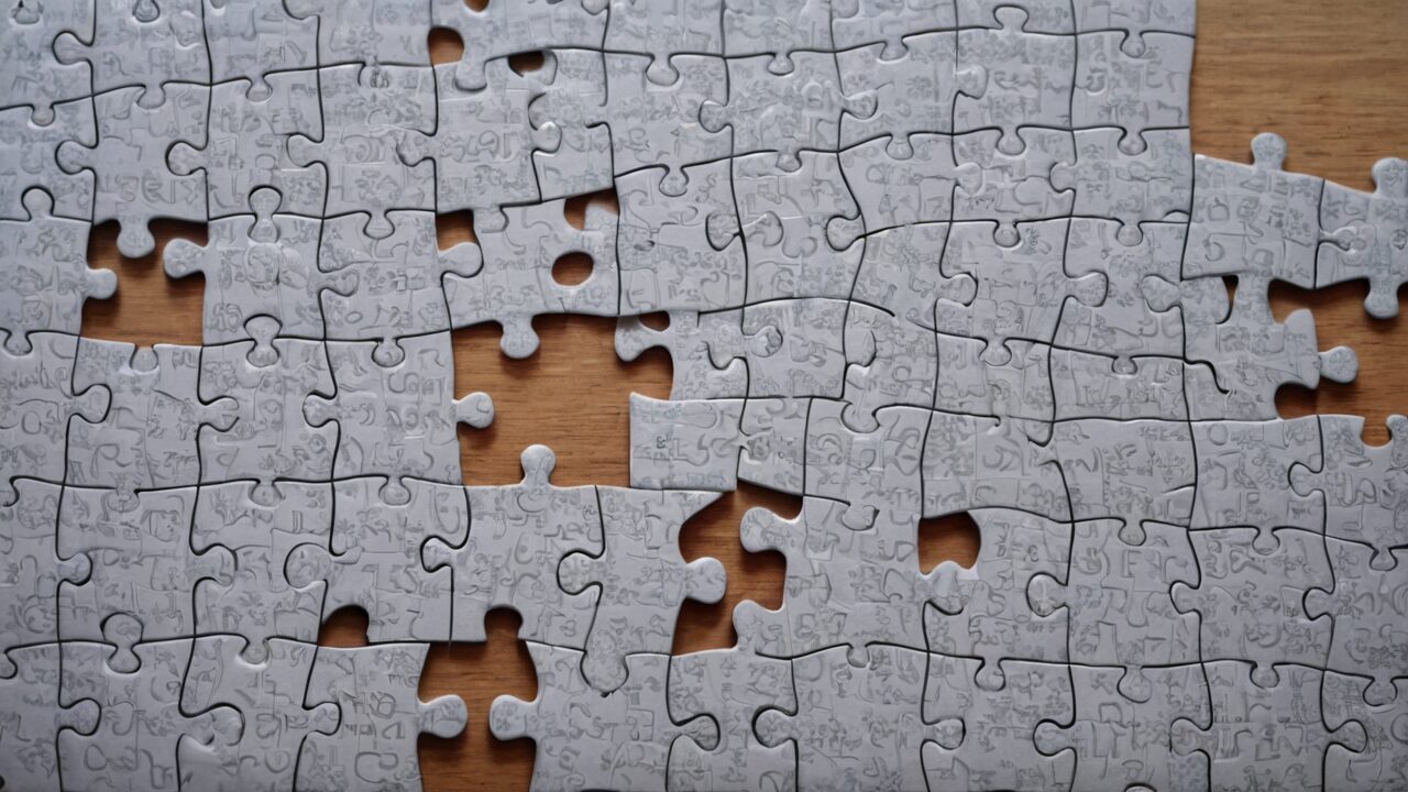 A partially completed jigsaw puzzle with many pieces missing, spread out on a wooden surface. The puzzle pieces are grey with various writings and doodles.